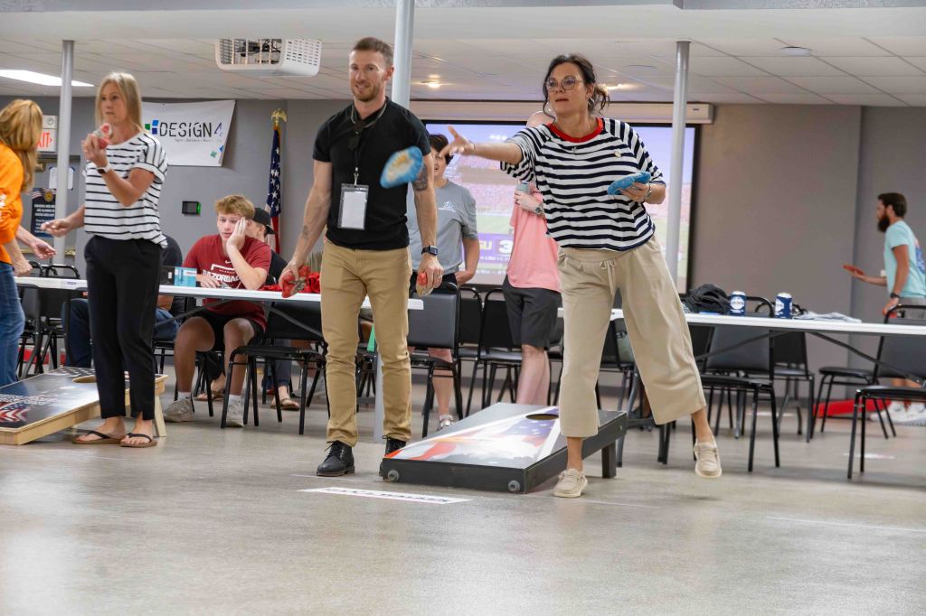 A group of people playing a bean bag toss game indoors, with one woman in a striped shirt throwing a bag towards the target.