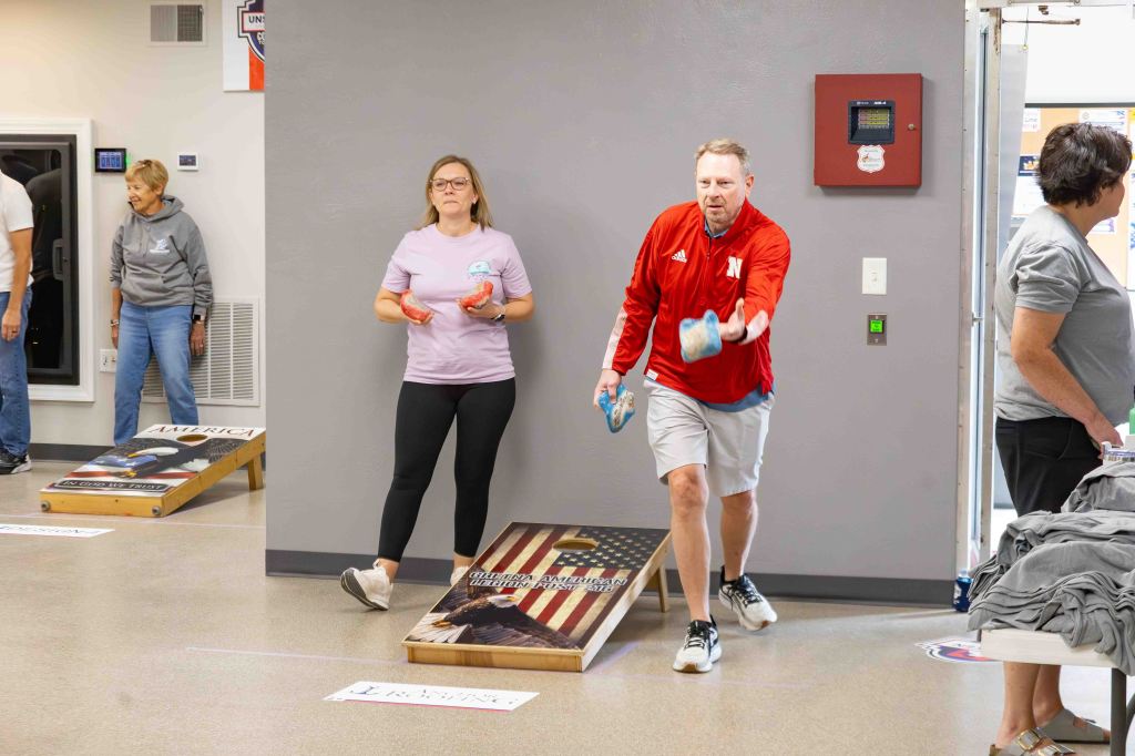A group of people playing cornhole indoors, with one person throwing a blue bean bag towards a wooden board featuring an American flag. Another woman stands ready to toss a red bean bag, while two others are observing the game. The setting is casual with t-shirts and a stack of shirts nearby.