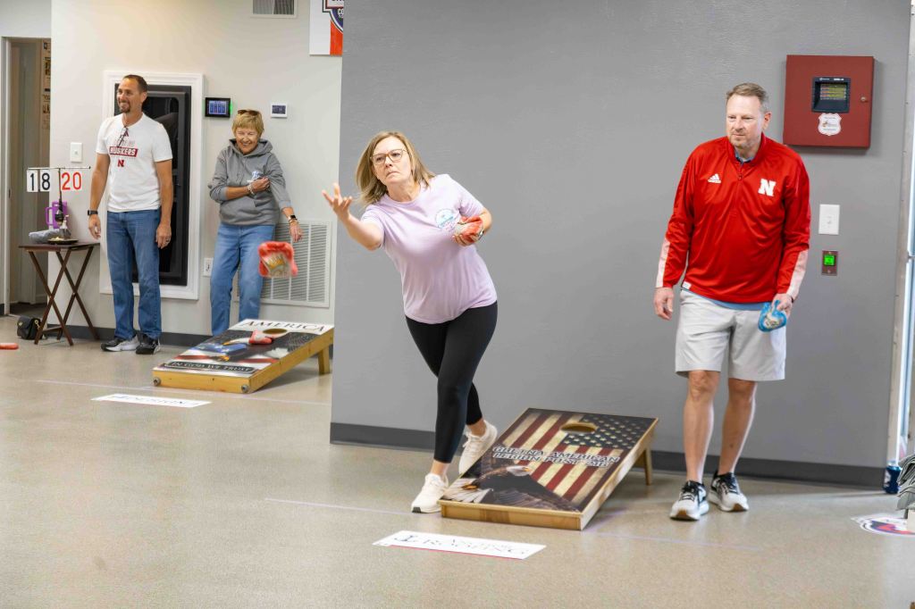 A group of people participating in a cornhole game inside a recreational facility. A woman in the foreground is tossing a bean bag while wearing a casual outfit. Two spectators are positioned in the background, with one holding a bean bag and the other observing the game. The cornhole boards feature a patriotic design.