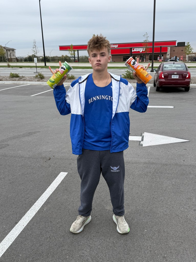 A young person standing in a parking lot, wearing a blue shirt and light jacket, holding a green Pringles can in one hand and an orange Pringles can in the other.