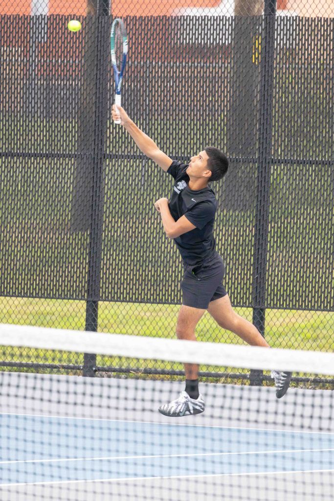 A young male tennis player serves the ball while standing on a court, with a fence in the background.
