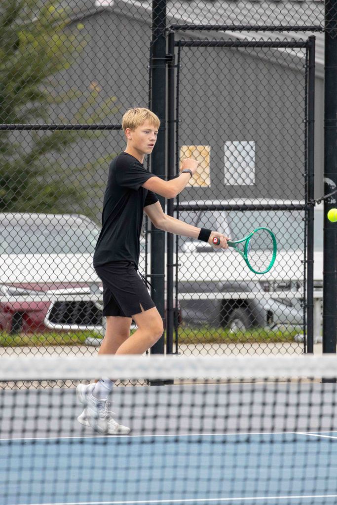 Young male tennis player in a black shirt and shorts, preparing to hit a tennis ball on a court.