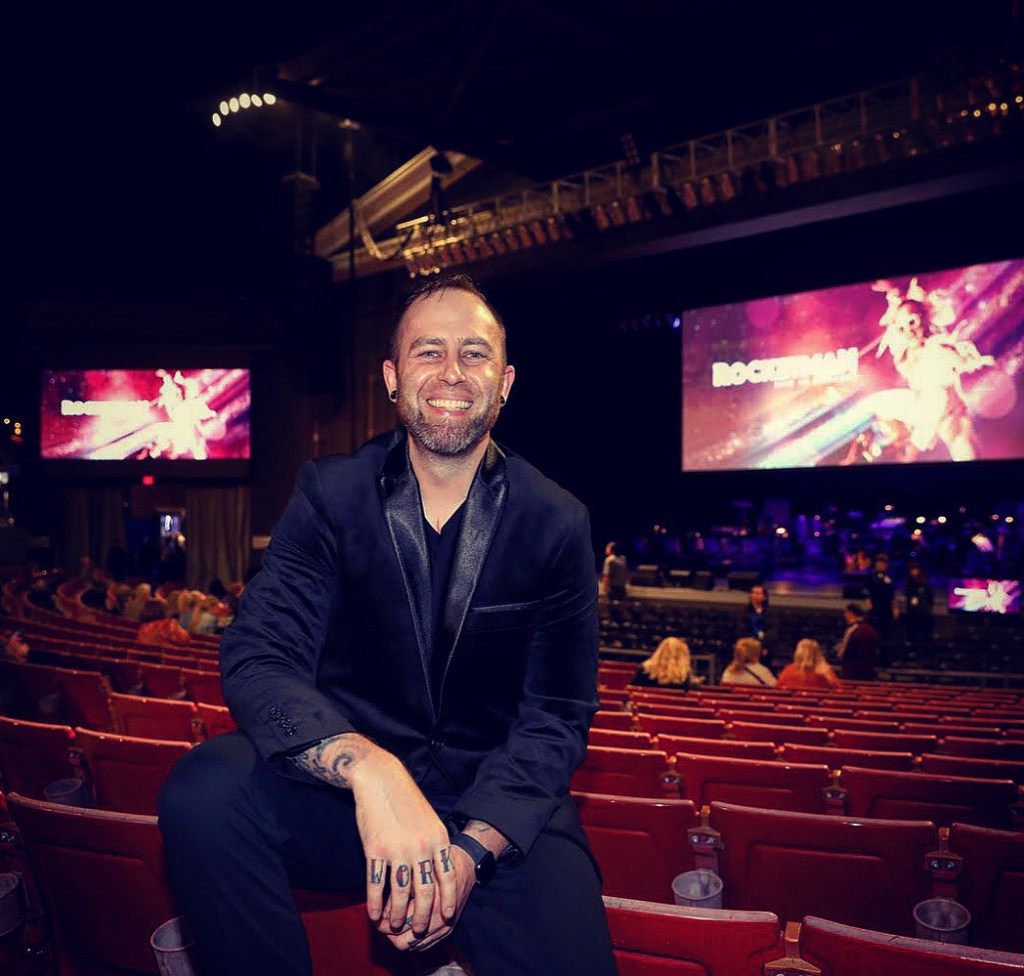Man sitting on a theater seat, smiling, wearing a black blazer. A large screen in the background displays a colorful announcement for 'ROCKETMAN' with an audience visible.