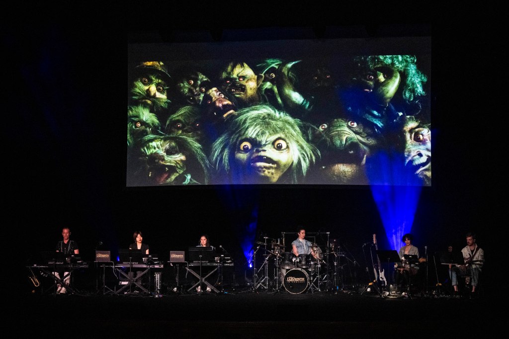 A live band performing on stage with keyboards and drums, accompanied by a large screen displaying various colorful creature faces.