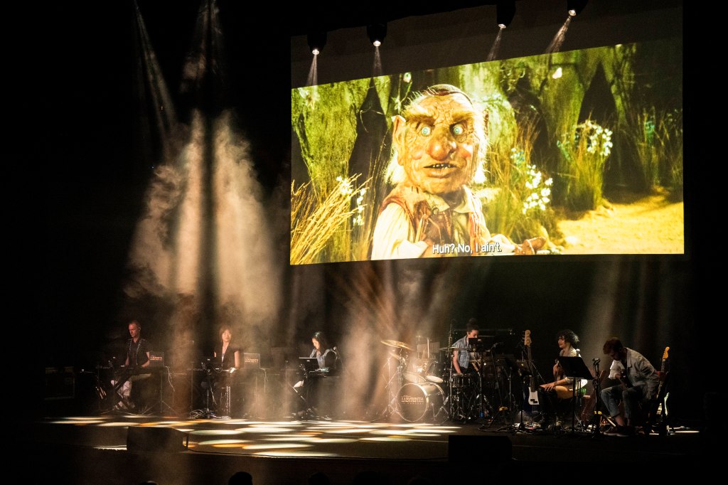 A live performance featuring musicians playing instruments on stage, with a large screen displaying a scene from a film featuring a puppet character.