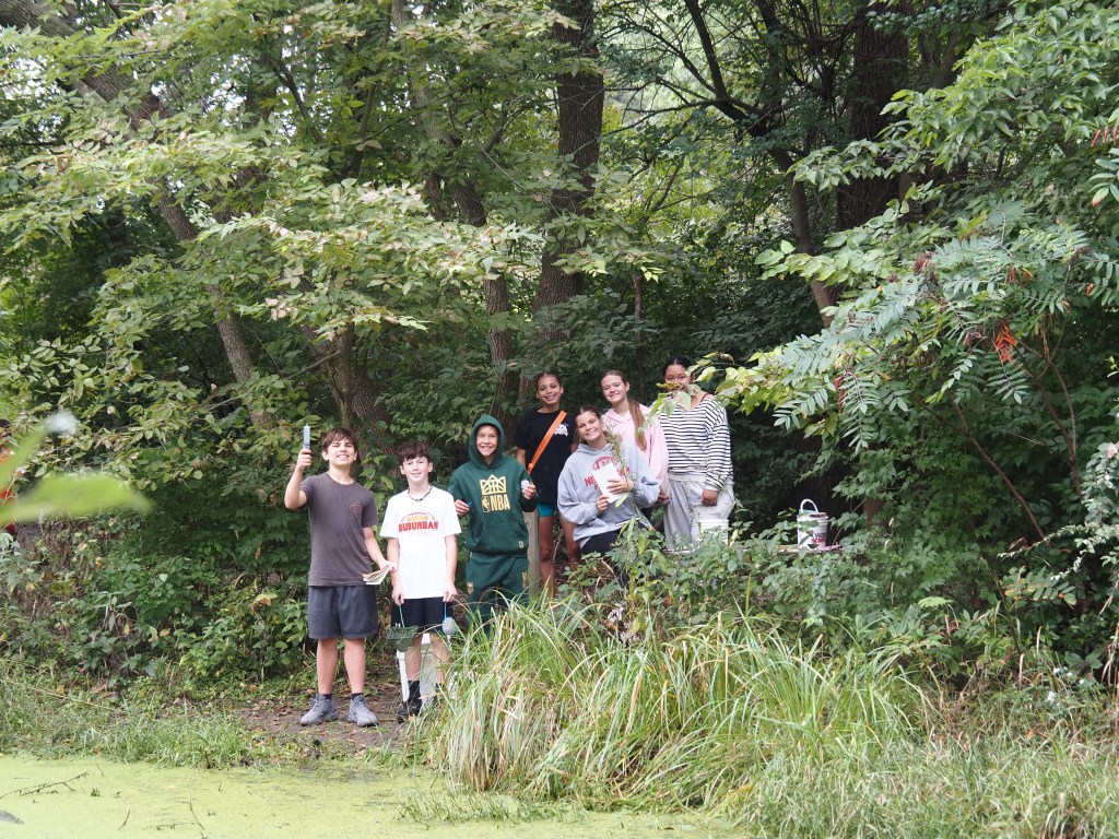A group of seven kids standing by a lush green area with trees, smiling and posing together. Some are holding objects and wearing casual clothing, while a pond with green vegetation is visible in the foreground.