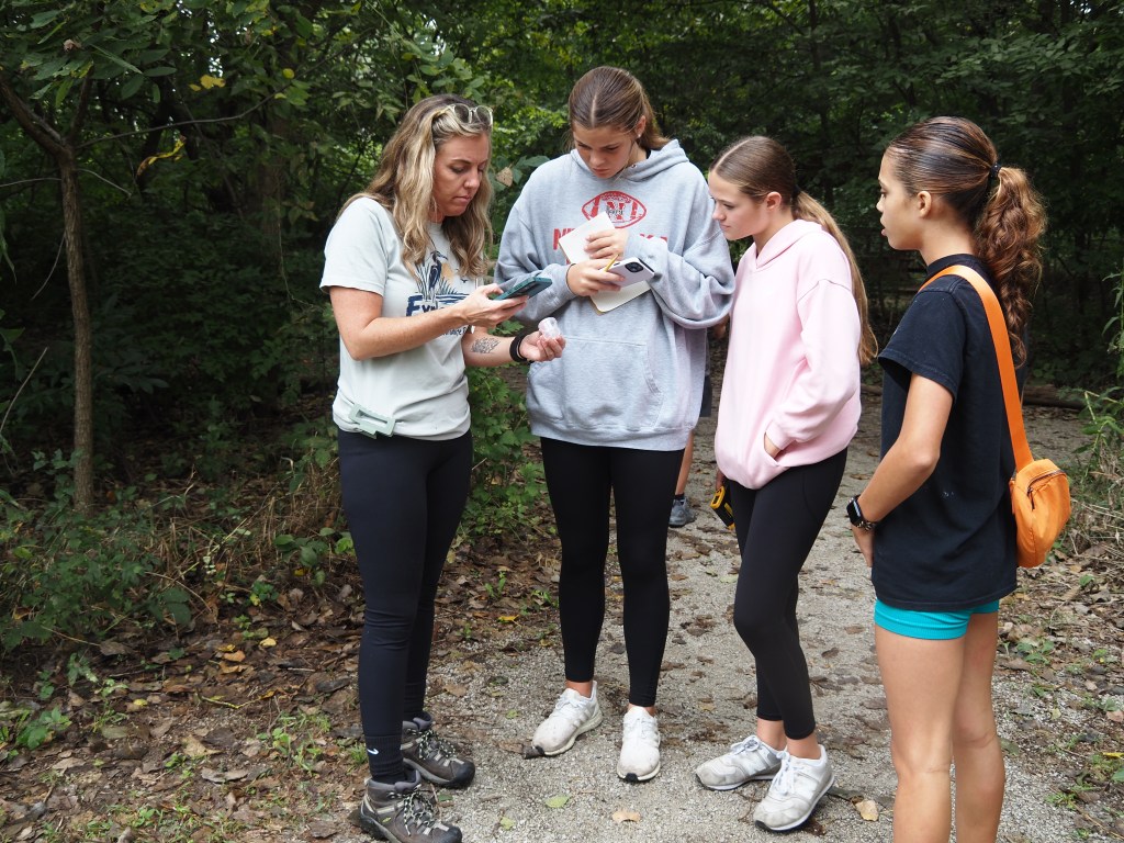 A group of four young women standing on a forest path, one woman demonstrating something on her phone to the others, who are observing attentively.