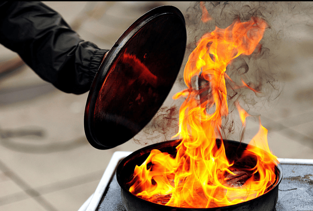 A person lifting a lid off a pot with flames and smoke erupting from inside.