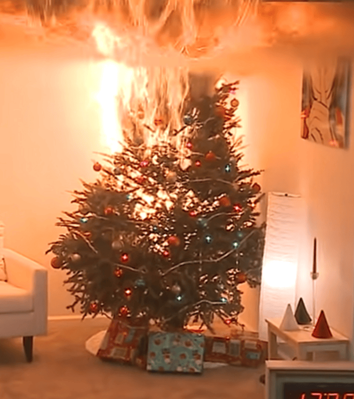 A Christmas tree engulfed in flames in a living room, with presents beneath the tree and a clock displaying the time.