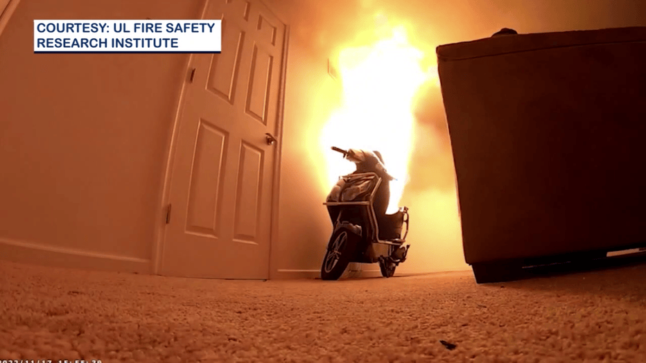 An electric scooter is engulfed in flames inside a room, with a couch partially visible in the foreground.